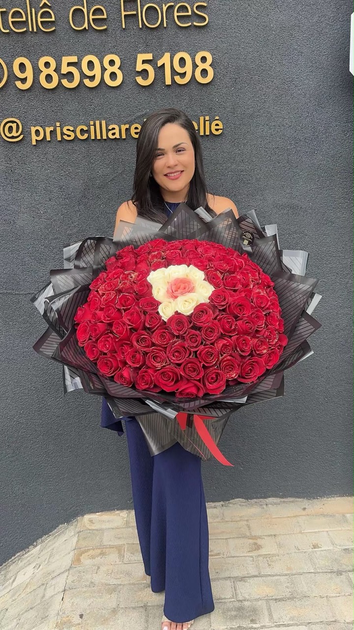 Priscilla diante do ateliê com um buquê circular de rosas vermelhas e flores claras no centro.